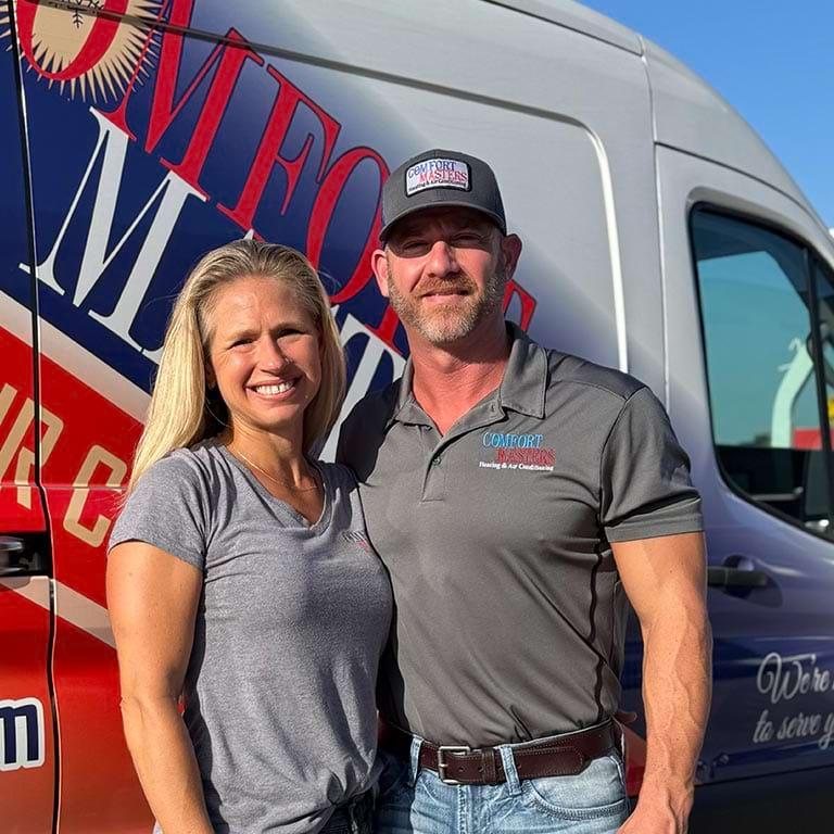 A smiling man and woman stand together in front of a white service van with a colorful Comfort Masters logo. Both are wearing casual clothes, and the man is also wearing a branded cap. Its a sunny day.